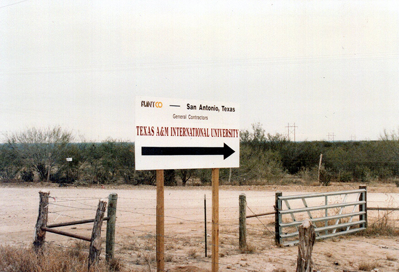 a plain white Contruction sign that reads 'Texas A&M International University' with a black arrow point to the right. Behind the sign is cleared out land.